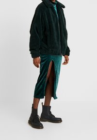 Chaqueta de forro polar verde con cuello, combinada con una falda de terciopelo verde azulado con una abertura lateral y medias de red negras, finalizando con botas negras con cordones.