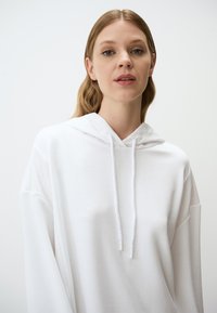 Biała, oversize'owa bluza z kapturem i kieszenią kangurką, z szyjącym sznurkiem kapturem i prążkowanymi mankietami, wykonana z miękkiego materiału mieszanki bawełny.