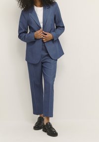Costume bleu marine ajusté avec une texture lisse, comprenant un blazer à simple boutonnage et un pantalon droit, assorti à des mocassins noirs.