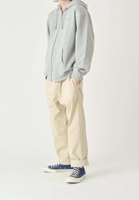 Grå dragkedjetröja med framficka, beiga vida byxor och blå sneakers med vita sulor. Bakgrunden är ljus och minimalistisk.