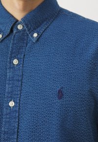 Blauw gestippeld overhemd met knopen, gemaakt van gestructureerde stof, met een kraag, witte knopen en een klein geborduurd logo op de borst.