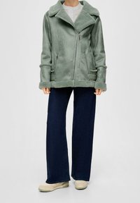 Persoon in een lichtgroene schapenvachtjack met een diagonale rits, donkere wijde spijkerbroek en beige sneakers tegen een effen achtergrond.