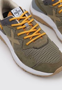 Zapatillas deportivas verdes y grises con tejido de malla, suela de goma, cordones naranjas y detalles de costura notables. Acentuadas con un logo y patrones.