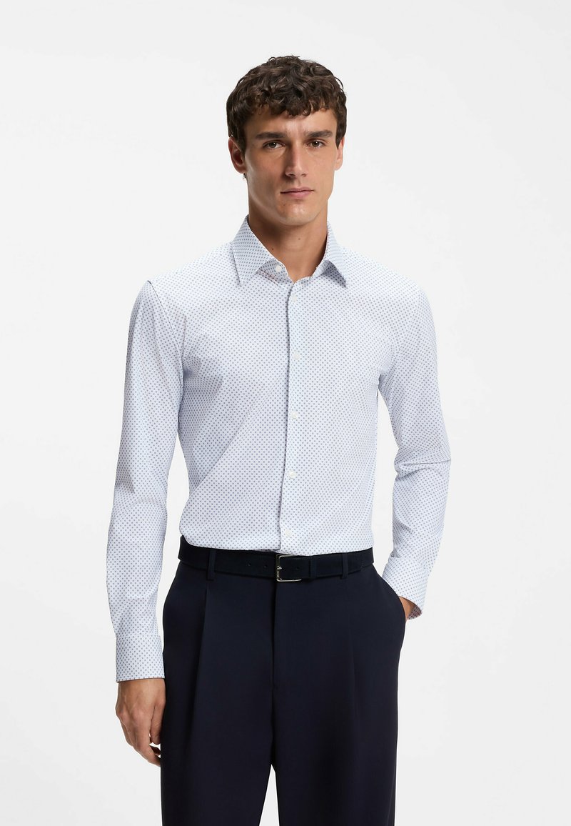 Homme portant une chemise blanche à manches longues avec de petits points bleus et un pantalon noir, debout avec une main dans la poche, sur fond blanc.