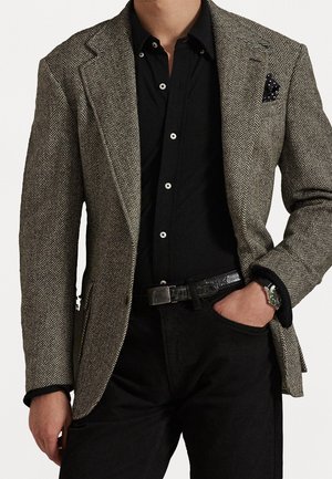 Homme portant un blazer à motif chevrons sur une chemise noire boutonnée et un pantalon noir, avec une main dans la poche et une montre-bracelet visible.