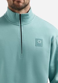 Lichtblauwe fleece pullover met een hoge kraag, halve rits en een vierkante patch met geometrische patronen op de linkerborst.