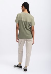 T-shirt verde oliva con un design a blocchi di colore, maniche corte e una texture liscia. Abbinato a pantaloni beige e scarpe nere con accenti gialli.