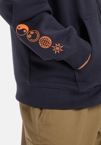 Navy blue zip-up hoodie featuring orange graphic prints of various celestial and geometric shapes on the left sleeve. Cotton blend fabric.