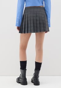 Persona con una mini gonna plissettata grigia, calze nere, stivali neri stringati e una camicia a righe blu a maniche lunghe, in piedi contro un muro bianco.