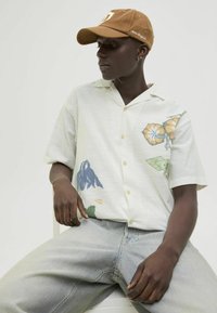 Short-sleeve button-up shirt with floral patterns in blue and yellow on a light background, paired with light gray denim jeans and a brown cap.