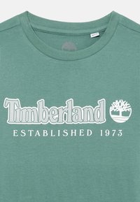 Grønn bomull T-skjorte med "Timberland"-logo og treikon i hvitt, med teksten "ETABLERT 1973" nedenfor. Rund hals-design.