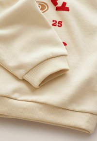 Cremefarbener Sweatshirtärmel mit geripptem Bund. Enthält rot- und braunbedruckten Text und Zahlen, weiche Textur und lockere Passform.