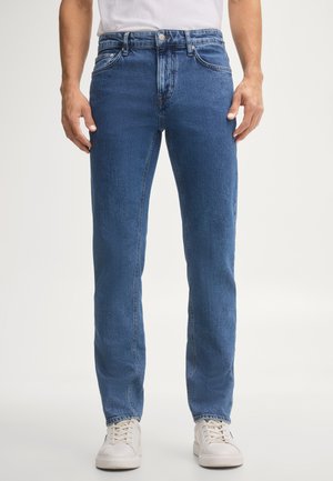 Mann trägt mittelblaue Jeans mit geradem Bein, weiße Sneakers und ein weißes T-Shirt, steht vor einem schlichten hellen Hintergrund.