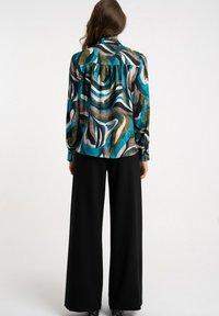 Kobieta stojąca tyłem, ubrana w bluzkę z długim rękawem o abstrakcyjnym wzorze w odcieniach niebieskiego, zielonego i beżowego, szerokie czarne spodnie oraz botki za kostkę.