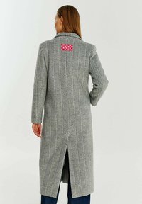 Šedý kabát ve vzoru herringbone s rozparkem na zadní straně, obsahující červenobílé kostkované aplikace. Střih na míru s klopovým límcem.