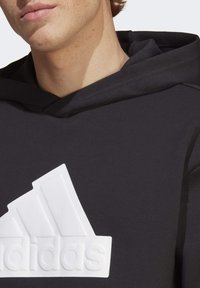 Black hoodie made of soft fabric, featuring a large white Adidas logo with a raised texture and three diagonal stripes.