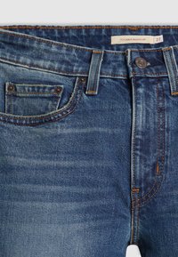 Jean en denim bleu avec bouton et fermeture éclair, poche avant, passants de ceinture, coutures orange et étiquette indiquant "725 Kick Bootcut 27" à l’intérieur de la ceinture.