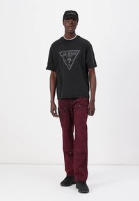 Sort bomulds t-shirt med et grått trekantet "GUESS" logo, parret med dyb røde baggy denim jeans og sorte sneakers. Simpelt design og teksturer.