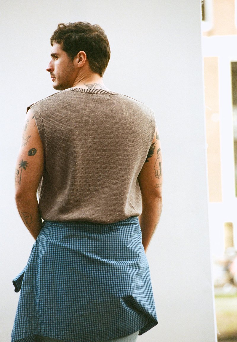 Muž s tetováním nosící béžový svetřík bez rukávů a modrou kostkovanou košili uvázanou kolem pasu, který se dívá stranou proti bílá stěně.