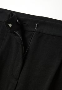 Zwarte broek met een soepel stof, ritssluiting, platte voorkant en een knoopsluiting. De tailleband heeft een lus en een riemgebied.