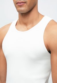 Top de malha branco com nervuras, com um decote redondo e alças largas, apresentando um logótipo em relevo subtil na frente.