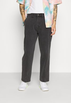 Homme portant un pantalon noir ample, des baskets blanches, une chemise blanche et une chemise à manches courtes colorée pastel, debout avec les mains dans les poches.