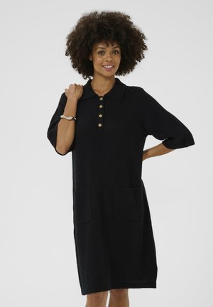 Woman with curly hair wearing a black knee-length dress with gold buttons, elbow-length sleeves, two front pockets, and a silver bracelet.