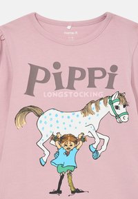 Roza majica s natpisom "Pipi Duga Čarapa" i ilustracijom djevojčice koja podiže bijelog konja s plavim točkama i zelenim akcentima.