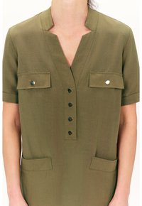 Tunic corta a maniche corte di colore verde oliva con scollatura a V, dettagli con bottoni, due tasche frontali e una texture liscia. Finiture argentate sui bottoni.
