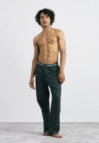 Calças verdes de corte solto com um cós elástico preto apresentando o texto "BJÖRN BORG". Tecido suave e design minimalista. Modelo de pé descalço.