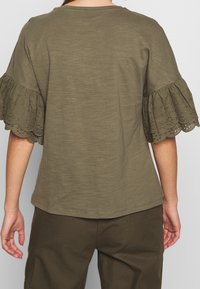 Femme portant un haut vert olive avec des manches courtes à volants brodés et un pantalon assorti vert olive, vue de dos.