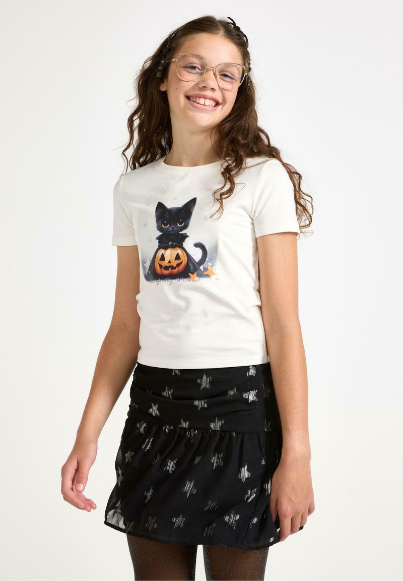 Valkoinen lyhythihainen t-paita, jossa on musta kissa ja kurpitsagrafiikka; yhdistettynä mustaan röyhelöskorttiin, jossa on hopeisia tähtikuvioita.