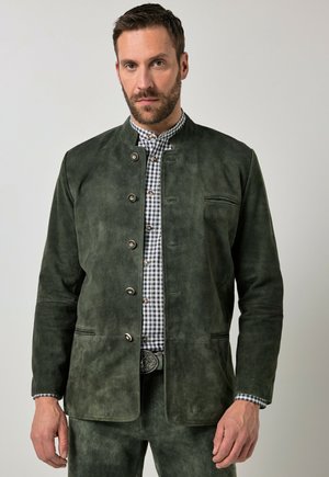 Mand iført mørkegrøn ruskindjakke over sort-hvid ternet skjorte, stående mod en ensartet lys baggrund.