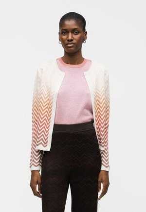 Kvinde iført en pink top og en hvid cardigan med orange zigzag-mønstre, kombineret med mørke high-waist bukser med zigzag-mønster.