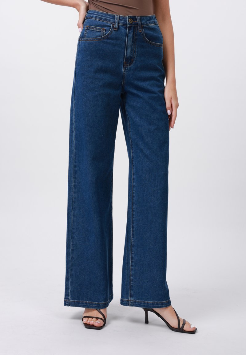 Plati džinsai tamsiai mėlynos spalvos, su aukštu liemeniu, matomomis siūlėmis, priekinėmis kišenėmis ir laisvu siluetu.