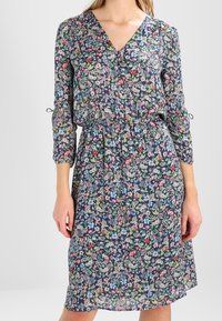 Kukkamekko syvällä V-pääntiellä, kootulla vyötäröllä ja kolmen neljänneksen hihoilla. Taustalla tummansininen väri, jossa on värikkäitä kukkakuoseja.