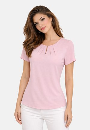 Kobieta o długich brązowych włosach, nosząca złote kolczyki-koła, jasnoróżową bluzkę z krótkim rękawem z małym wycięciem z przodu oraz białe spodnie.