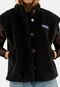 Zwarte fleece vest met een hoge kraag, knoopsluiting en twee voorkruiden. Heeft een klein kleurrijk logo-opstrijkertje op de borst.