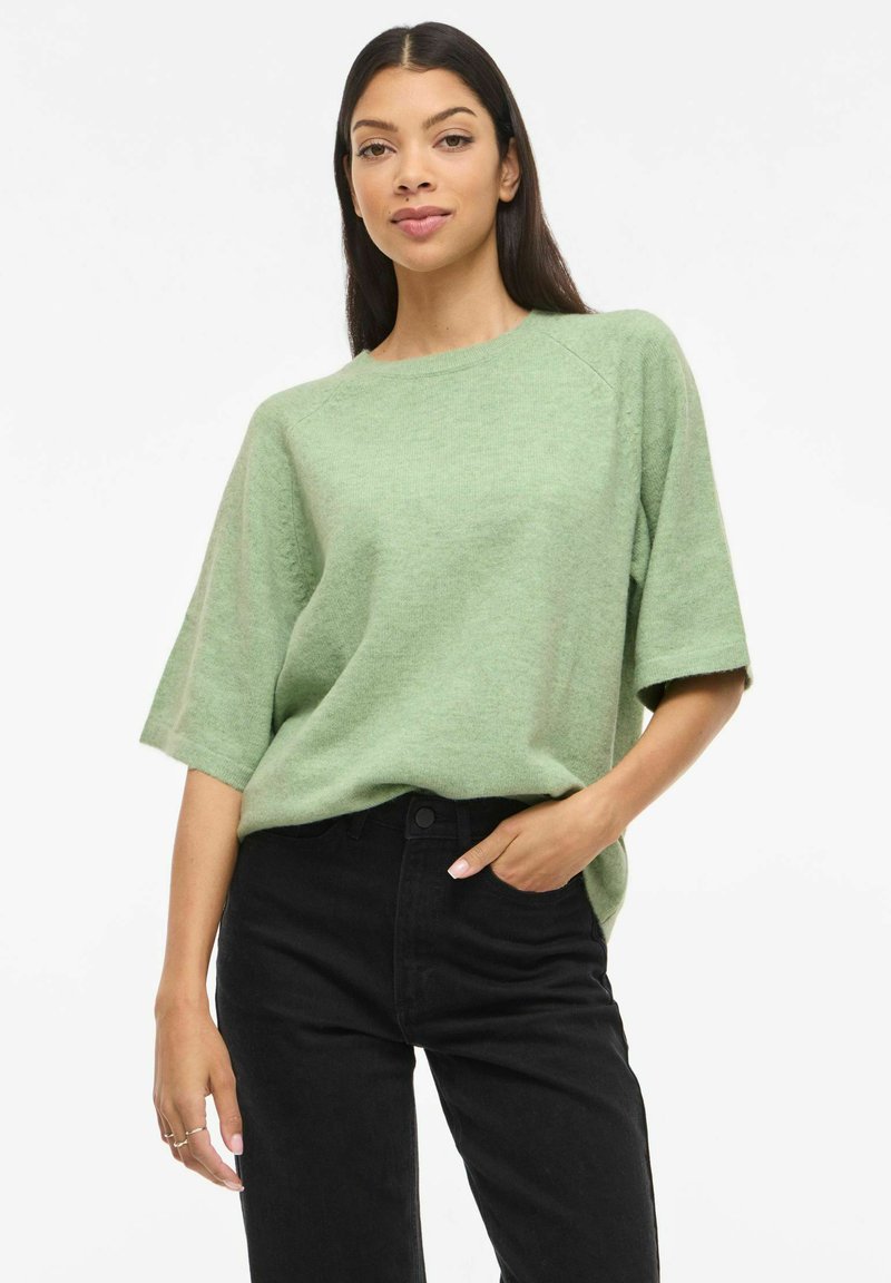 Suéter de punto verde menta con mangas cortas, cuello redondo y ajuste relajado, combinado con jeans negros. Textura suave, detalles sutiles en las costuras.