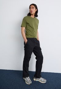 Een jonge man staat in een casual houding, gekleed in een olijfgroen T-shirt en zwarte cargo-broeken, met zijn handen in zijn zakken tegen een neutrale achtergrond.