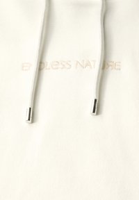 Witte hoodie met een geribbelde textuur, voorzien van de geborduurde frase "ENDLESS NATURE" in een lichte beige kleur. Zilverkleurige hardware op de trekkoorden.
