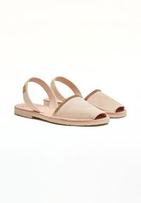 Sandalias de ante beige con la punta abierta, suela plana y tiras traseras en el tobillo. Presentan un detalle metálico decorativo en la parte superior.