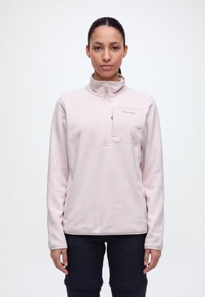 Une femme porte une polaire Columbia rose pâle à fermeture éclair courte, avec une poche poitrine et un pantalon noir, sur un fond clair uni.