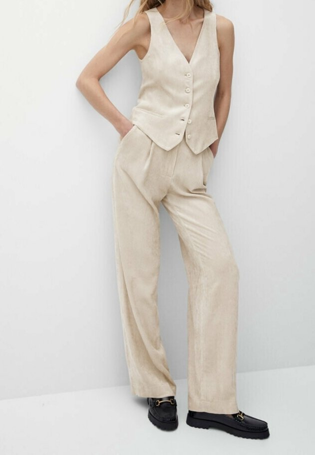 Femme portant un gilet beige sans manches boutonné et un pantalon large assorti avec des mocassins noirs, debout les mains dans les poches.