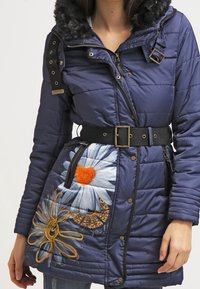 Chaquetón azul marino con capucha de pelo sintético, bordados florales en azul y naranja, cintura ajustada con un cinturón negro, cierres de cremallera y botón. 