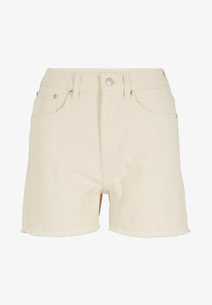 Persona vestida con una sudadera con capucha color crema, pantalones cortos a juego con dobladillos sin rematar y zapatillas altas blancas, de pie en un entorno interior neutro.