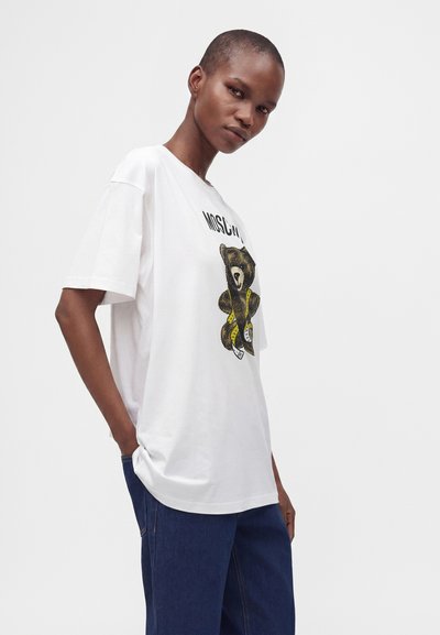Άτομο που φοράει λευκό φαρδύ T-shirt με σχέδιο αρκούδας και μπλε τζιν, στέκεται με το ένα χέρι στην τσέπη σε μονόχρωμο φόντο.