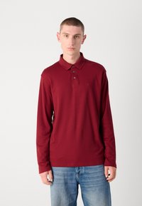 Burgundi színű, hosszúujjú polo ing, puha anyagból készült. Klasszikus gallérral, három gombos zárással és egy kis logóval rendelkezik a mellkas részén.