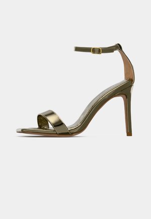 Gouden hoge hak sandaal met enkelbandje en gesp, open teen, slanke stilettohak, en glanzende metalen afwerking op een effen achtergrond.