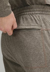 Ruskeat urheiluhousut, joissa on teksturoitu kangas ja vetoketjullinen tasku lantiolla. Housuissa on joustava vyötärö ja kontrastiväriset ompeleet.